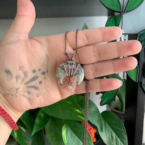May include: A handcrafted necklace featuring a round, patterned stone pendant wrapped in copper wire. A copper crescent moon and a wire spider with a pink bead accent the pendant. The necklace has a copper chain.