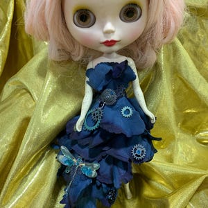 May include: A doll wearing a blue dress with a dragonfly and gears. The dress is made of fabric that looks like petals. The doll has pink hair and blue eyes.