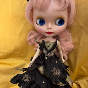 May include: A Blythe doll with pink hair and large blue eyes. The doll wears a black and green dress with petal-like layers and decorative accents. A black headpiece with green accents adorns the doll's head. The doll is posed against a yellow background.
