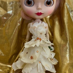 May include: A doll with large eyes, pink hair, and red lips, wearing a layered cream-colored dress adorned with small flowers and sparkling embellishments. The doll is set against a gold, textured background.