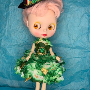 May include: A doll wearing a green and gold dress with a shamrock design. The doll has pink hair and is wearing a black top hat with a rainbow and shamrock embellishment. The doll is standing on a blue surface with gold coins scattered around.