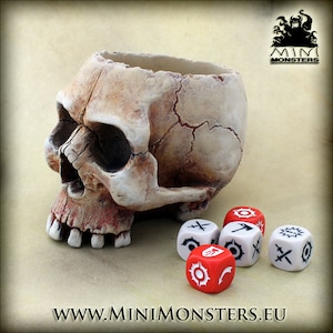 Könnte beinhalten: Ein schädelartiger Behälter mit verwittertem, knochenähnlichem Aussehen. Mehrere Würfel mit weißen und roten Flächen, die jeweils einzigartige Symbole zeigen, sind darum herum angeordnet. Die Website-Adresse www.minimonsters.eu ist unten sichtbar.