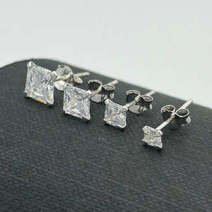 Puede incluir: Cuatro pares de pendientes de botón cuadrados de plata con piedras de circonita cúbica transparentes. Los pendientes están ordenados por tamaño, con el par más grande a la izquierda y el par más pequeño a la derecha.