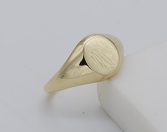 May include: A gold signet ring featuring a smooth band and a textured oval face. The ring has a classic design and a polished finish. The oval face is approximately 1.3 cm in diameter. This ring is a piece of jewellery.