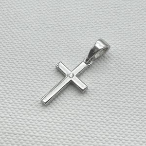 May include: A silver-colored pendant in the shape of a cross. The cross has a small, round, clear stone at the center. The pendant is designed with a loop at the top for a chain. The background is a textured, off-white surface.