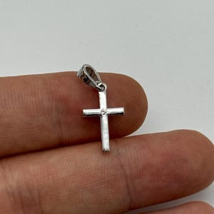 May include: A small, silver-colored cross pendant with a single, clear gemstone at the center. The cross has a simple, classic design and is suspended from a small loop. The pendant is likely made of metal and is shown against a neutral background.