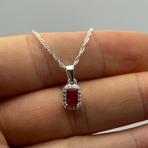 Puede incluir: Un collar de plata con un colgante rectangular de color rojo rubí. El colgante está rodeado por un halo de pequeñas piedras transparentes. La delicada cadena de plata realza el diseño elegante del collar.