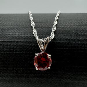 Könnte beinhalten: Eine silberne Halskette mit einem runden, tiefroten Edelsteinanhänger. Der Anhänger wird von vier Krappen gehalten. Die Kette hat ein filigranes, gedrehtes Design. Die Halskette wird auf einer schwarzen Oberfläche präsentiert.