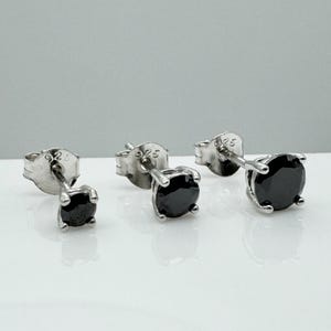 May include: Three pairs of stud earrings with black gemstones set in silver. The earrings have a classic, minimalist design, with a round black stone held by four prongs. The posts are straight and the backs are butterfly style.