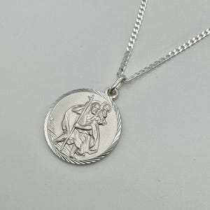 May include: A silver-colored St. Christopher pendant necklace. The round pendant features a detailed relief of St. Christopher carrying a child, with a textured border. The chain is a delicate curb style.