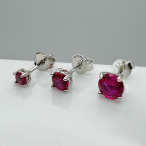 Può includere: Tre orecchini a lobo in argento con pietre preziose rotonde e rosso vivo. Gli orecchini sono esposti su una superficie bianca, mettendo in risalto il loro metallo lucido e il colore intenso delle pietre. Gli orecchini sono un classico della gioielleria.