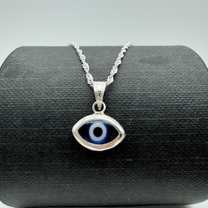 Puede incluir: Un collar plateado con un colgante en forma de ojo con un dise&ntilde;o azul y blanco. El colgante cuelga de una delicada cadena. El collar se exhibe sobre una superficie negra sobre un fondo blanco.