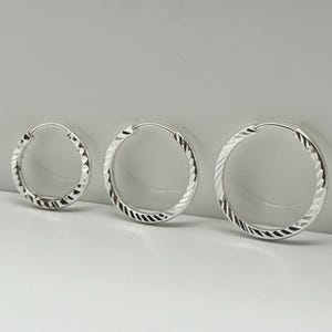 Könnte beinhalten: Drei silberne Creolen in verschiedenen Größen. Jede Creole hat ein strukturiertes, geriffeltes Muster am äußeren Rand. Die Ohrringe werden auf einer weißen Oberfläche vor einem weichen, neutralen Hintergrund präsentiert.