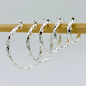 May include: Four silver hoop earrings with a twisted design. The earrings are displayed on a wooden surface.