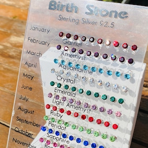 May include: A display of sterling silver birthstone stud earrings. The clear acrylic display lists months and corresponding gemstones like garnet, amethyst, aquamarine, crystal, emerald, ruby, sapphire, and topaz. The text "Birth Stone" is at the top.