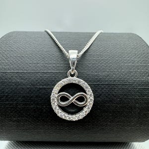 Puede incluir: Un collar plateado con un colgante con el símbolo del infinito. El colgante presenta un círculo de pequeñas piedras transparentes que rodean el símbolo del infinito. La cadena es una fina cadena de cable plateada.