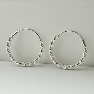 May include: A pair of silver hoop earrings with a twisted rope design. The earrings are circular and have a shiny, metallic finish. The hoops are shown against a neutral background.