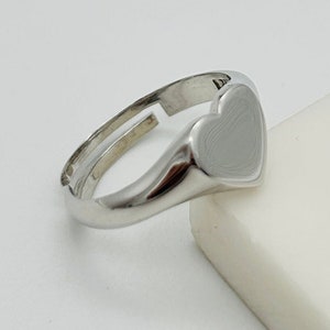 Könnte beinhalten: Ein silberner Ring mit einem herzförmigen Design. Das Herz ist glatt und hat eine leicht strukturierte Oberfläche.