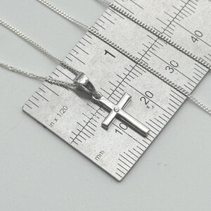 May include: A silver-colored cross pendant necklace with a small gemstone at the center of the cross. The necklace is on a silver chain, and a ruler is in the background. The cross measures approximately 0.5 inches in length.