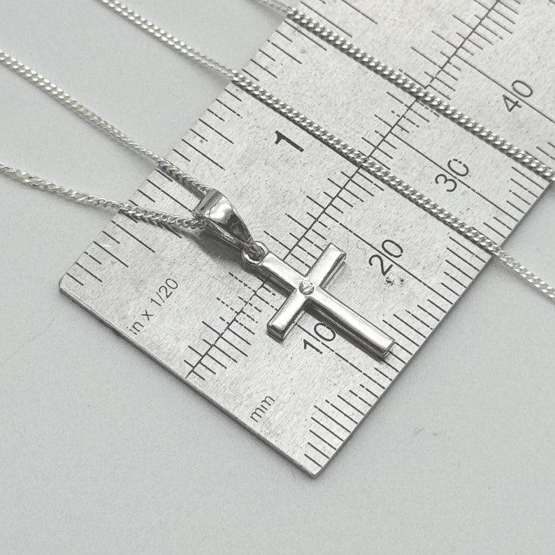 May include: A silver-colored cross pendant necklace with a small gemstone at the center of the cross. The necklace is on a silver chain, and a ruler is in the background. The cross measures approximately 0.5 inches in length.