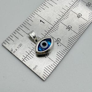 Puede incluir: Un colgante plateado con forma de ojo, con un dise&ntilde;o azul y blanco. El colgante mide aproximadamente 2 cm de largo y se muestra junto a una regla para la escala. El dise&ntilde;o del ojo tiene una pupila negra central.
