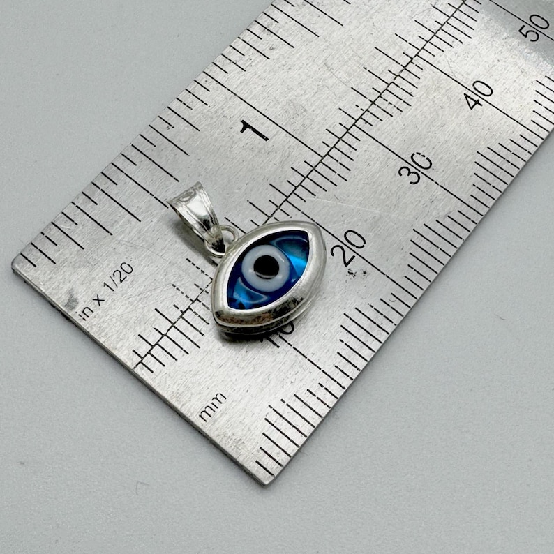 Puede incluir: Un colgante plateado con forma de ojo, con un dise&ntilde;o azul y blanco. El colgante mide aproximadamente 2 cm de largo y se muestra junto a una regla para la escala. El dise&ntilde;o del ojo tiene una pupila negra central.