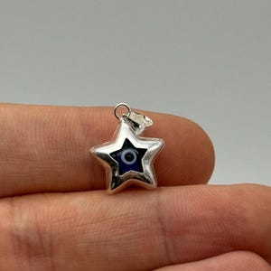 May include: A silver star pendant featuring a blue and white eye design. The pendant has a small loop for a chain. The star is a five-pointed shape. The pendant is likely made of a metal such as silver.
