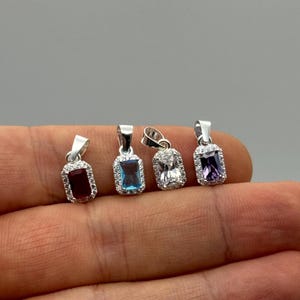 Puede incluir: Cuatro colgantes rectangulares de piedras preciosas con engastes plateados. Las piedras son rojas, azules, transparentes y moradas, cada una rodeada por un halo de piedras más pequeñas y transparentes. Los colgantes se muestran sobre un fondo neutro.