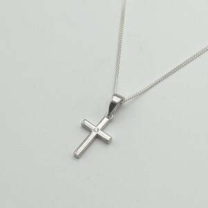 May include: A silver cross pendant necklace. The cross is a simple, classic design with a small, round accent in the center. The chain is delicate and silver, complementing the pendant. The necklace is displayed against a plain white background.
