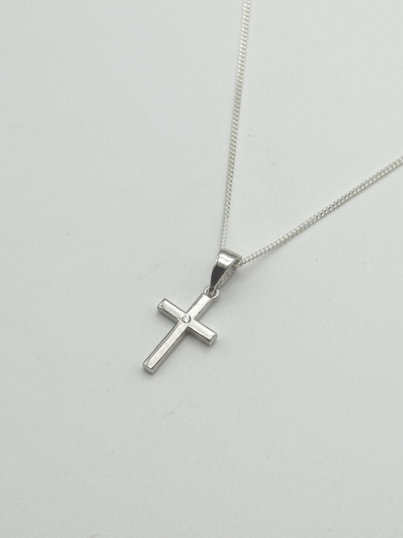 May include: A silver cross pendant necklace. The cross is a simple, classic design with a small, round accent in the center. The chain is delicate and silver, complementing the pendant. The necklace is displayed against a plain white background.