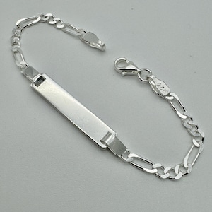 Peut inclure: Un bracelet en argent avec une plaque nominative rectangulaire et un design de chaîne Figaro. Le bracelet est en métal brillant et poli et comporte une fermeture à fermoir mousqueton. Le bracelet est présenté sur un fond gris clair.