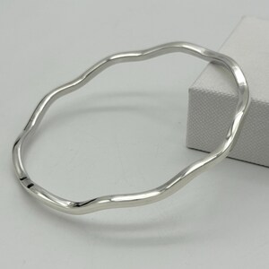 Puede incluir: Un brazalete plateado con un diseño ondulado y orgánico. El brazalete tiene un acabado liso y pulido y un estilo sencillo y elegante. El brazalete es un bucle continuo.
