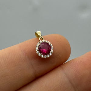May include: A gold pendant with a large, round, deep red gemstone surrounded by a halo of smaller, clear gemstones. The pendant has a small loop at the top for a chain. The jewellery is set against a neutral background.
