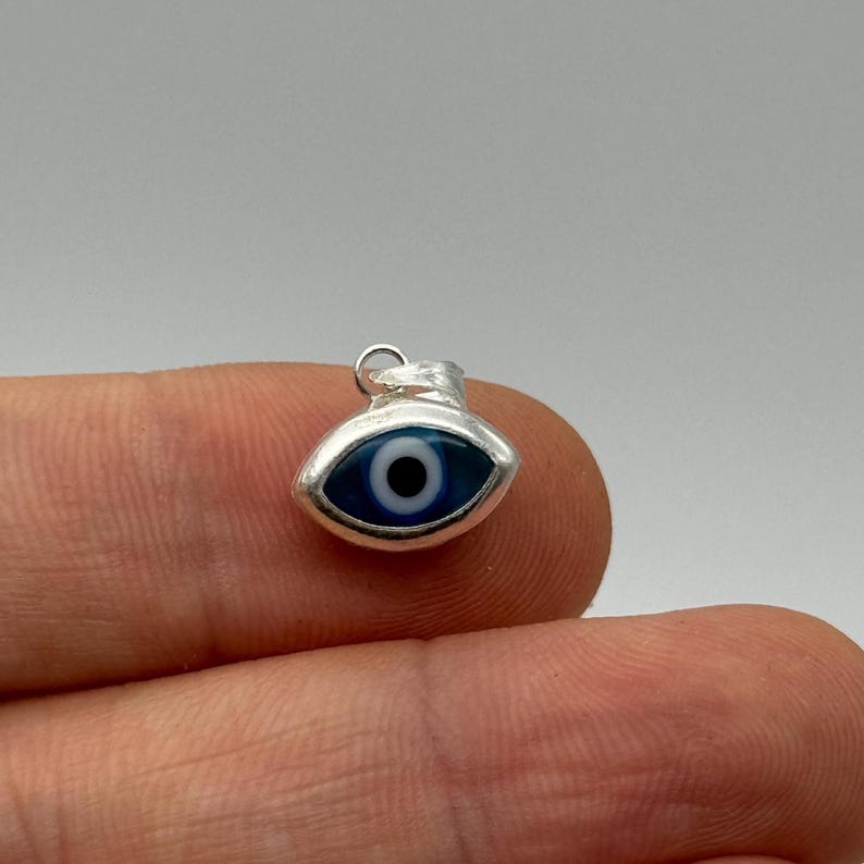 Puede incluir: Un peque&ntilde;o dije plateado con forma de ojo. El ojo presenta un iris azul oscuro con una pupila blanca y negra. El dije tiene un lazo en la parte superior para sujetarlo a un collar o pulsera.
