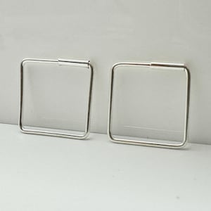 Könnte beinhalten: Ein Paar quadratische silberne Creolen. Jeder Ohrring hat ein quadratisches Design mit einem kleinen Steg oben. Die Ohrringe werden vor einem hellgrauen Hintergrund gezeigt.