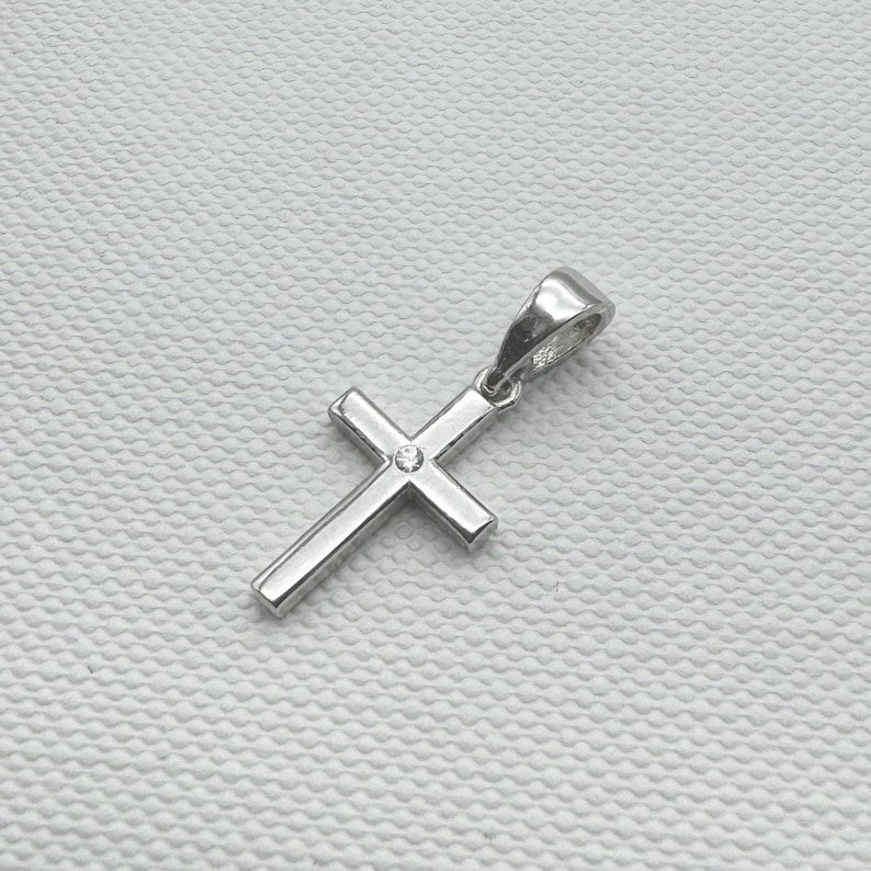 May include: A silver-colored pendant in the shape of a cross. The cross has a small, round, clear stone at the center. The pendant is designed with a loop at the top for a chain. The background is a textured, off-white surface.