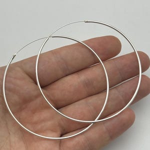 Peut inclure: Une paire de grandes créoles circulaires fines en argent. Les boucles d'oreilles présentent un design simple en fil continu avec une finition métallique lisse et polie. Chaque anneau est léger et conserve une forme parfaitement ronde.