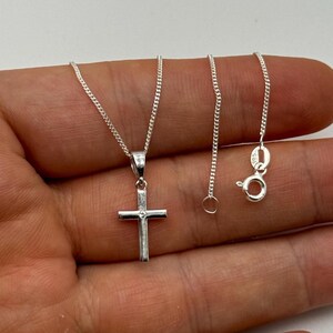 May include: A silver cross pendant necklace. The cross is a simple design with a small, round accent. The chain is a delicate, linked style. The necklace is displayed against a neutral background.