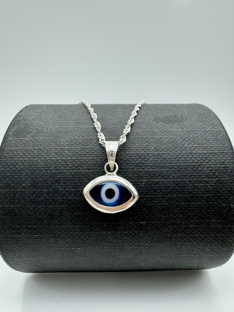 Puede incluir: Un collar plateado con un colgante en forma de ojo con un dise&ntilde;o azul y blanco. El colgante cuelga de una delicada cadena. El collar se exhibe sobre una superficie negra sobre un fondo blanco.