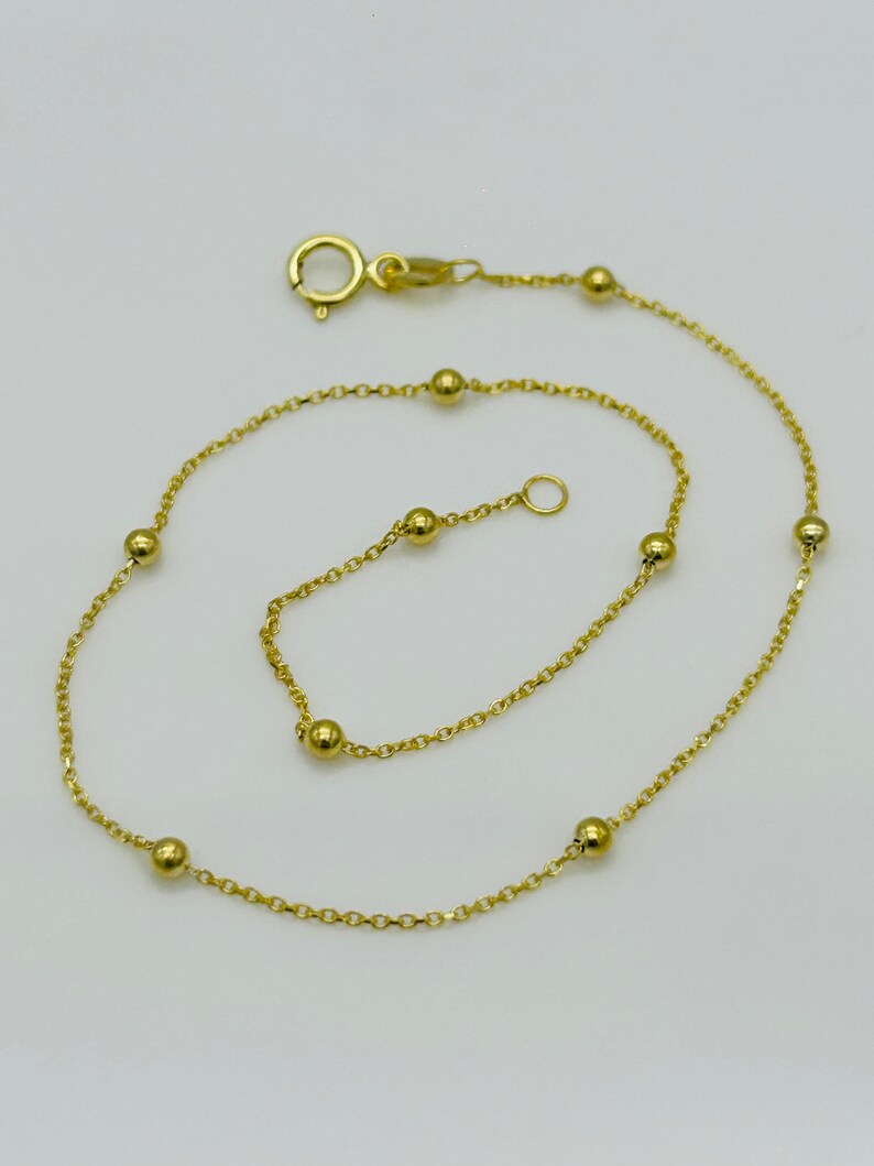 May include: A gold chain anklet with small round beads. The anklet has a lobster clasp closure.