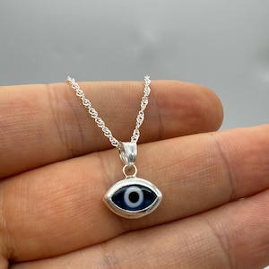 Puede incluir: Collar plateado con un colgante en forma de ojo. El colgante tiene un iris azul, una pupila negra y un reflejo blanco. La cadena es de dise&ntilde;o delicado y retorcido. El colgante mide aproximadamente 1,5 cm de largo.
