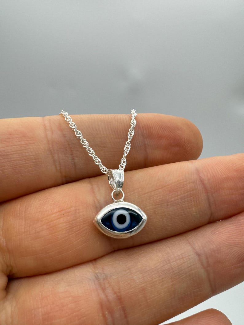Puede incluir: Collar plateado con un colgante en forma de ojo. El colgante tiene un iris azul, una pupila negra y un reflejo blanco. La cadena es de dise&ntilde;o delicado y retorcido. El colgante mide aproximadamente 1,5 cm de largo.