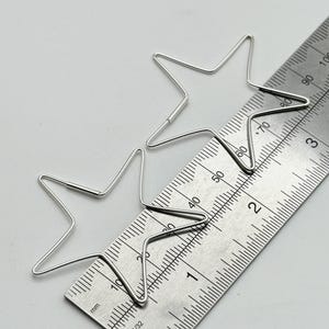 May include: Two silver wire star-shaped earrings. The earrings are next to a metal ruler with millimeter and inch markings. The stars are approximately 1.5 inches in diameter. The earrings are on a light gray surface.