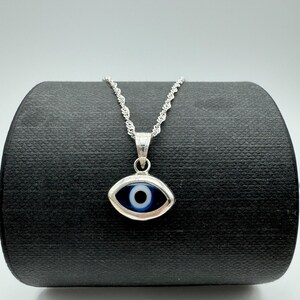 Puede incluir: Un collar plateado con un colgante en forma de ojo. El colgante tiene un dise&ntilde;o de ojo azul y blanco, engastado en un marco plateado. La cadena del collar es de estilo cuerda retorcida delicada, y el colgante est&aacute; suspendido de una peque&ntilde;a argolla.
