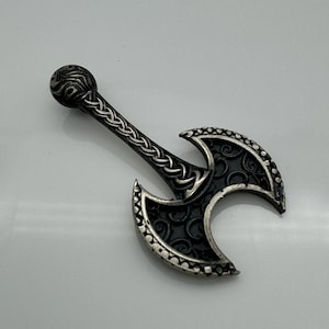 May include: A silver-coloured, double-bladed axe pendant. The handle features Celtic knotwork, and the blades have decorative patterns. The axe head is black with silver trim. The handle has a textured, rounded end.