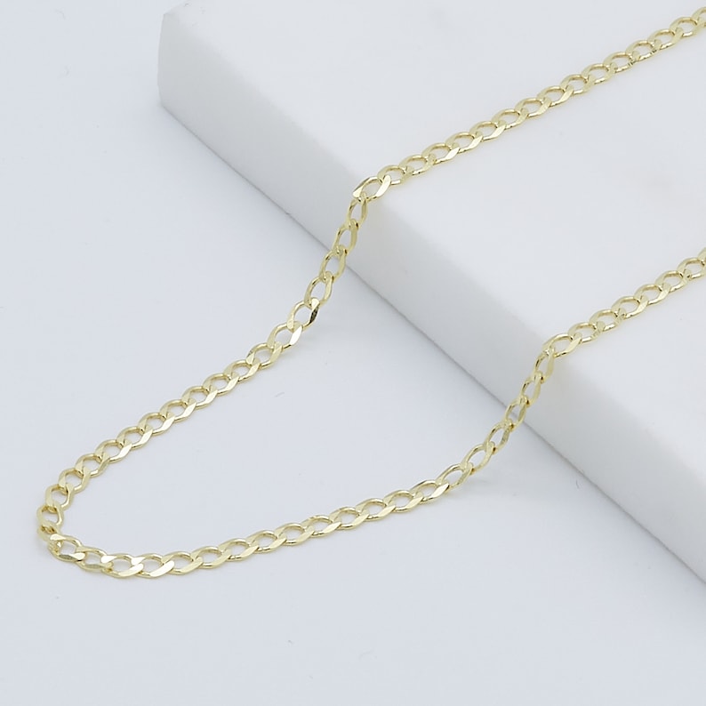 May include: A gold chain necklace with a curb link style. The chain is laying flat on a white surface.
