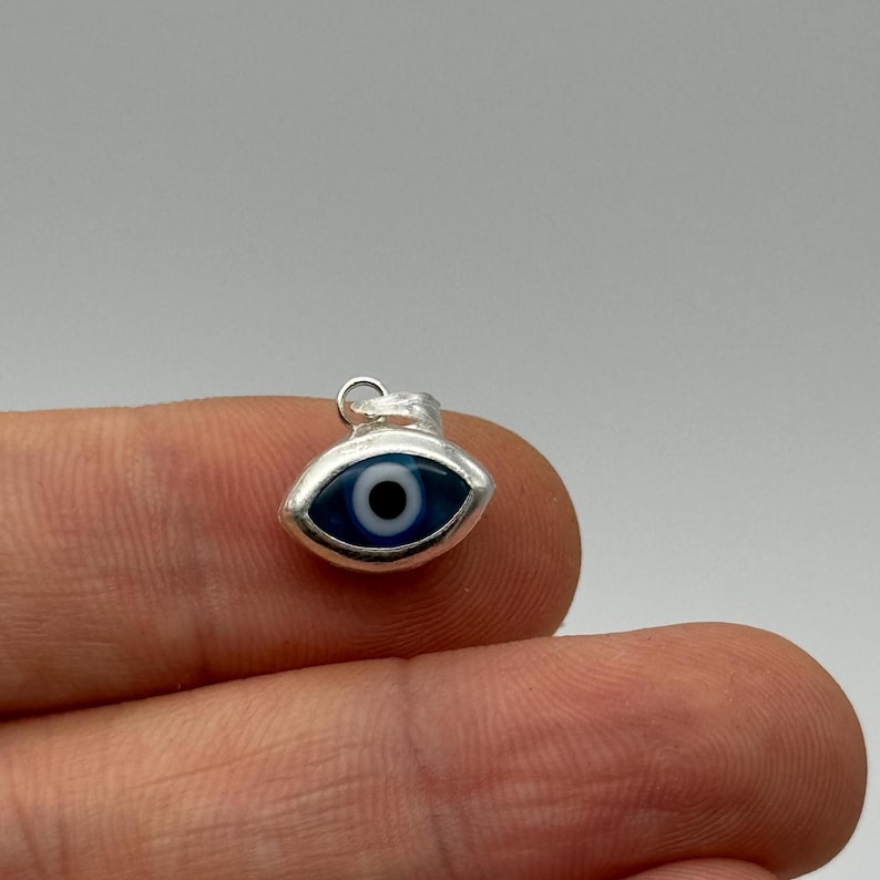 Puede incluir: Un peque&ntilde;o dije plateado con forma de ojo. El ojo presenta un iris azul oscuro con una pupila negra, sobre un fondo blanco. El dije se sostiene entre los dedos, mostrando su tama&ntilde;o y dise&ntilde;o. El dije tiene un peque&ntilde;o lazo en la parte superior para sujetarlo.