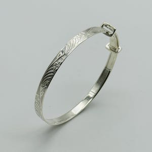 May include: A silver adjustable bangle bracelet with an engraved floral pattern. The bracelet has a clasp closure. The engraved design covers a portion of the bracelet's surface. The bracelet is displayed against a plain background.