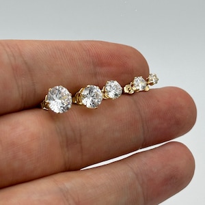 May include: A set of five gold stud earrings with clear round cubic zirconia stones. The earrings are arranged in order of size, with the largest earring on the left and the smallest earring on the right.