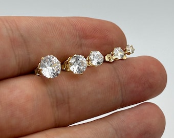 May include: A set of five gold stud earrings with clear round cubic zirconia stones. The earrings are arranged in order of size, with the largest earring on the left and the smallest earring on the right.